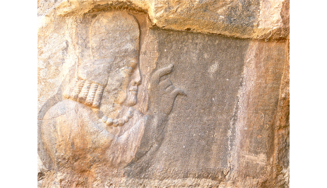A rock relief with the head and torso of a man on the left, and an inscription on the right. The man is raising his right index finger.