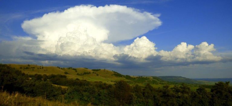 Photograph across green fields of large cumulus clouds against a blue sky with a huge cumulonimbus cloud, the top of which has spread into an anvil