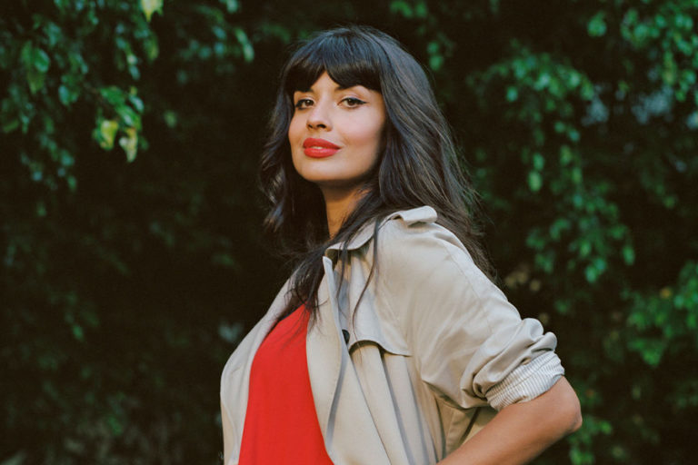 A photograph of Jameela Jamil wearing a light brown coat, red dress with matching red lipstick and long, dark brown hair. Jameela poses in front of some greenery