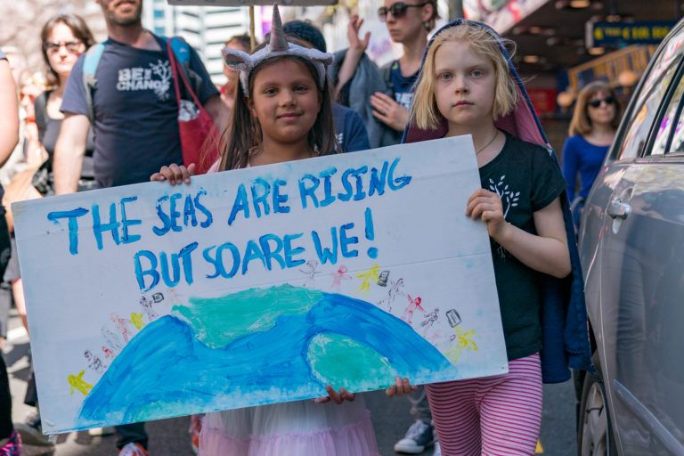 Two girls at a climate change protest holding a banner that has an image of the worlds with the writing "The seas are rising but so are we!"