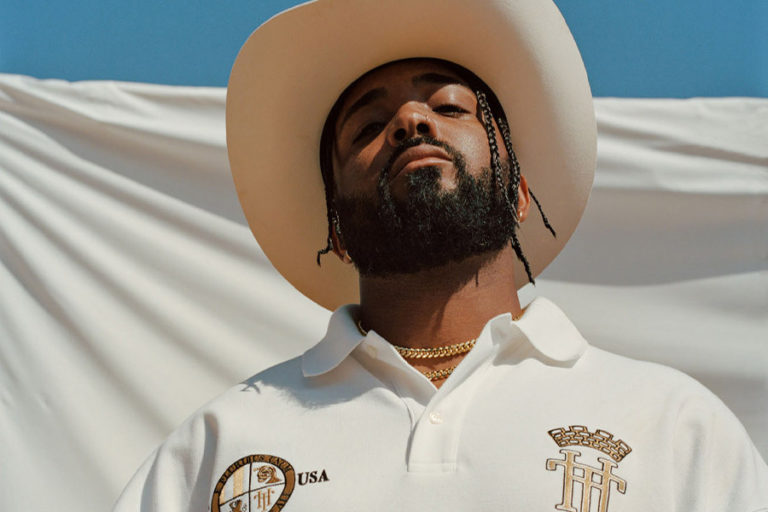 Randy stands, looking down at the camera with a white sheet behind him and blue sky in the background. He is wearing a white polo t-shirt and a cowboy hat.
