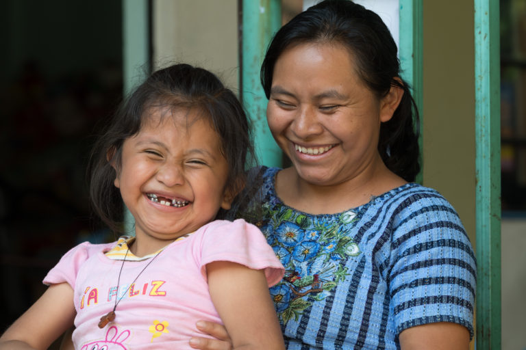 A girl is sitting on her mother's lap whilst they both laugh