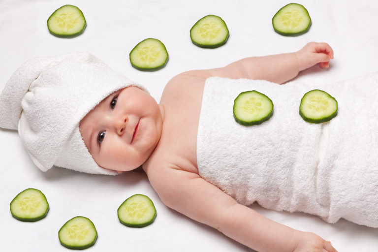 A comical image of a newborn baby wrapped in a towel. She lies back with a towel swaddled around her head as though she is in a beauty salon and there are pieces of cucumber all around her, as though she is in a spa environment