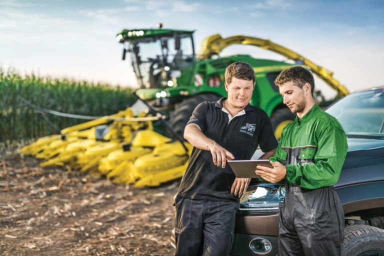 Two men in a field discussing something on a hand-held tablet with a large item of agricultural machinery behind them