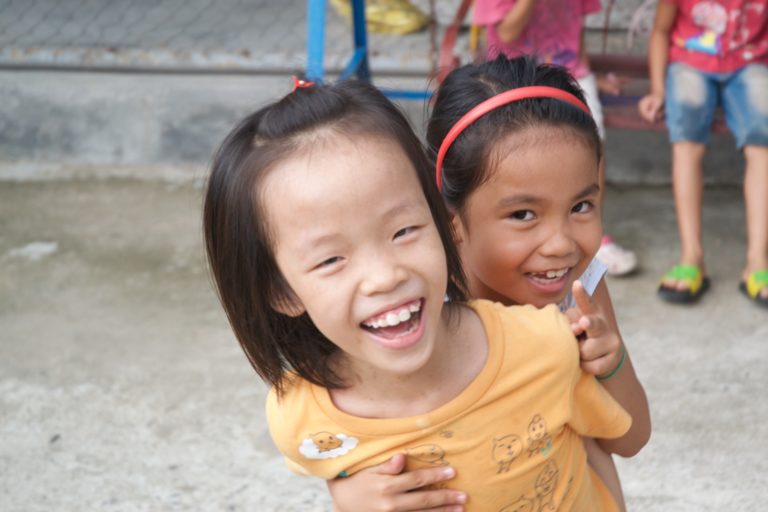 Two girls lean on each other and laugh. The girl on the left has a genetic syndrome