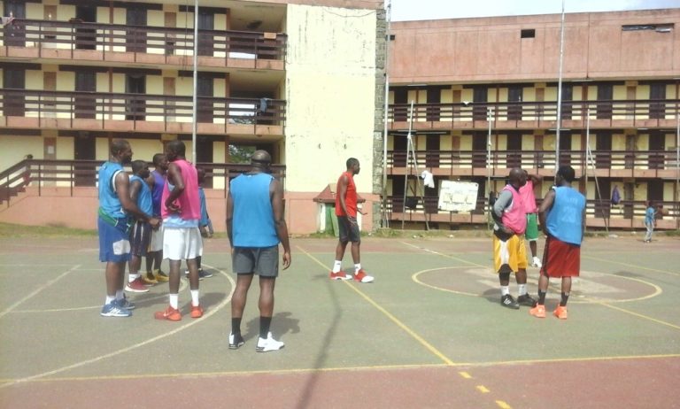 Group of men on a campus in the capital of Cameroon, dressed in sleeveless vests and shorts, gathered to play basketball together
