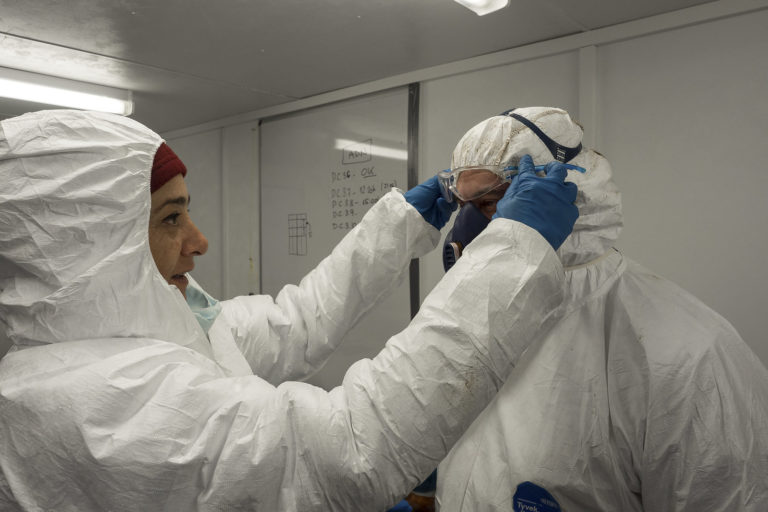 Two people are putting on white coveralls and masks to prevent contamination of the bodies with their own DNA during excavation