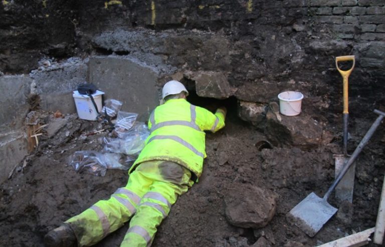 An archaeologist dressed in high visibility rainproof clothes lies on his front on piles of soil whilst reaching under a wall to excavate