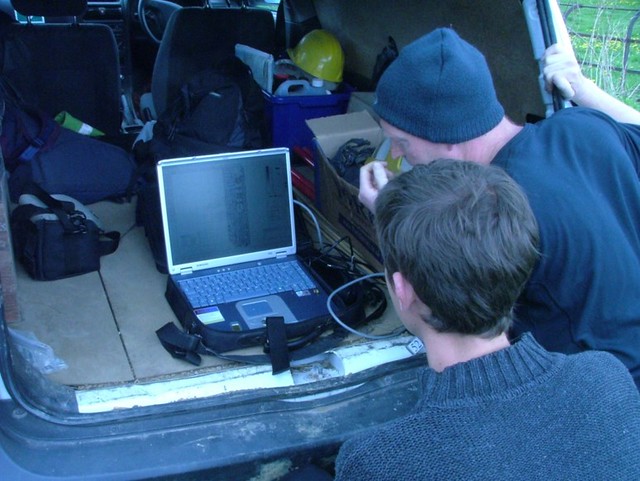 Two people looking at a laptop computer which is resting on the boot of a van. They are processing geophysical data