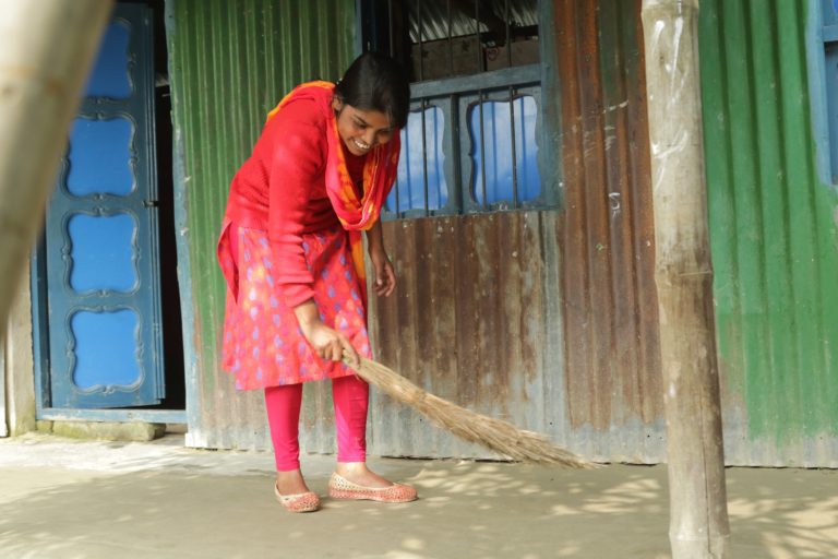 A teenage girl is sweeping outside her home as she smiles.