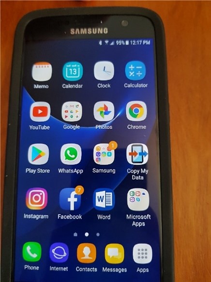 Fairly large photo of an Android smartphone's screen manufactured by Samsung, displaying a large number of apps on its home screen, including the calendar, clock, memo, Youtube, Google, WhatsApp, Facebook, Instagram, Microsoft Word, Calculator and Google Chrome browser amongst others. There are also apps in folders called 'Microsoft apps' and there is also a folder called 'Samsung'. At the bottom are icons for more 'basic' functions such as accessing one's list of contacts, messages, voice calling, and others.