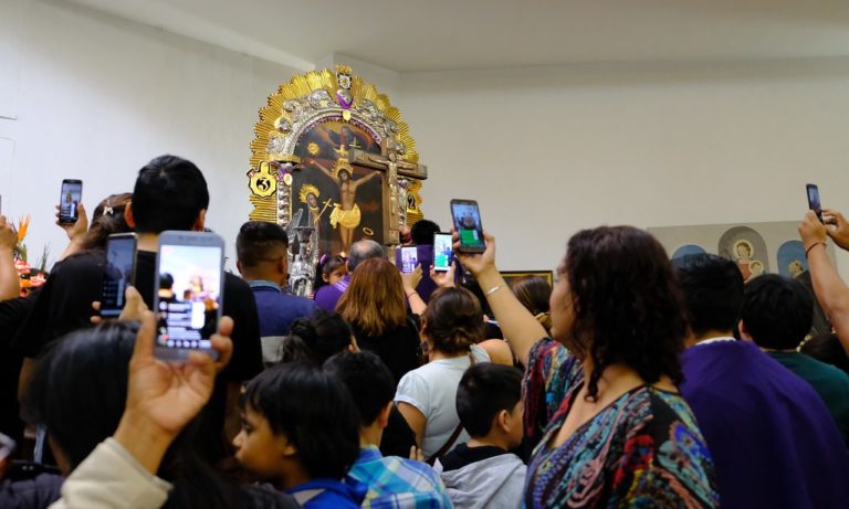 Interior of a church with a painting of the Christ, surrounded by people aiming their smartphones at the image. These are Peruvian migrants honouring in Chile the Lord of Miracles, the most revered Peruvian religious icon. The devotees broadcast with the smartphones the celebration to their friends and family abroad.