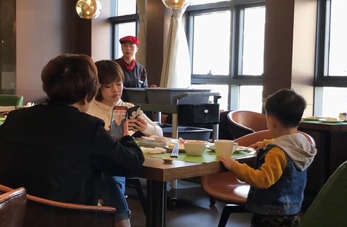 Two women, a younger and an older one are sitting at a restaurant, at a wooden table, and each looking down at their phones, one is editing a photo, next to them is a young toddler who is being ignored, there is also a waiter with a dark red cap transporting multiple trays of ingredients behind them