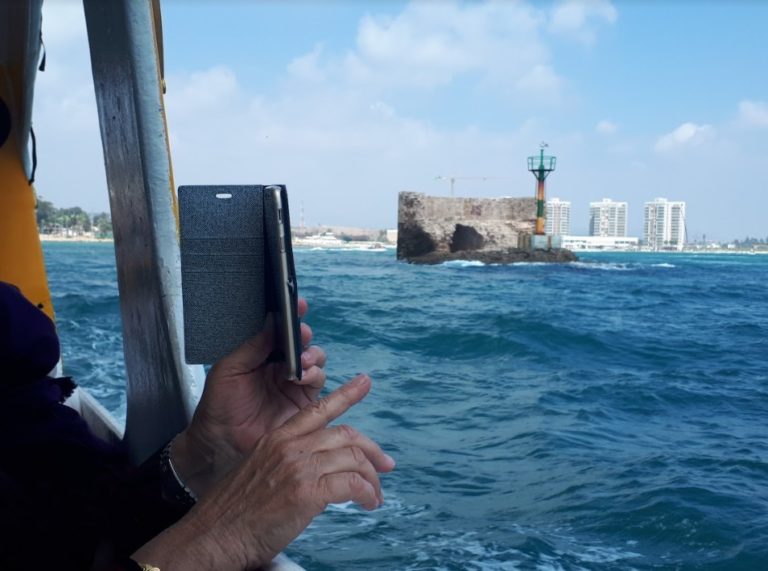 The hands of a woman are seen holding her smartphone as she takes a photo of something in the distance while she is on a boat, the blue sea can be seen around the boat