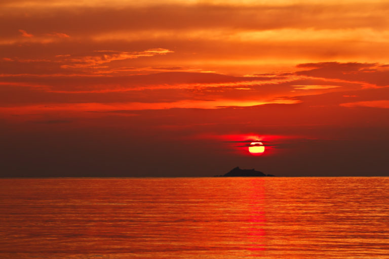 Sun setting over the sea with an island in the distance. The sky is different shades of red