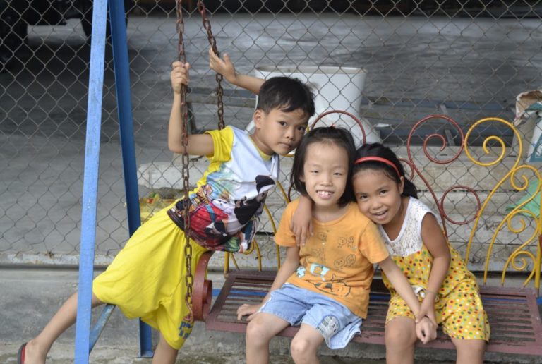 Two girls sitting side by side on a swing and smiling. A boy is standing next to the swing.