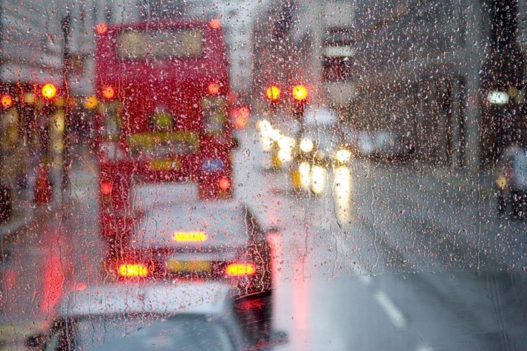 Photograph out through a rain-covered bus window in London looking at vehicles on a road at traffic lights in the rain