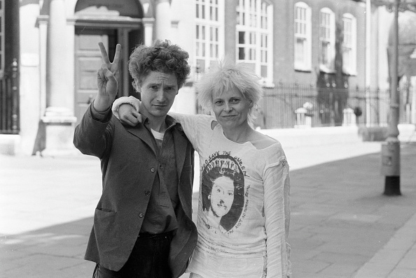 black and white photo. Malcolm is making a victory sign and Vivienne wears a shirt printed with the Sex Pistols' image of the Queen