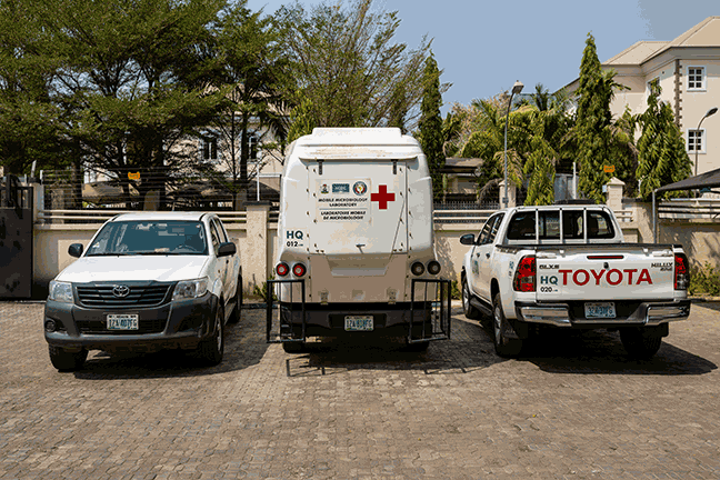 Three vehicles are stationed in a parking lot. These include two white pick-up trucks, one facing forward and one facing backward and a white mobile laboratory truck with a large red cross at its back