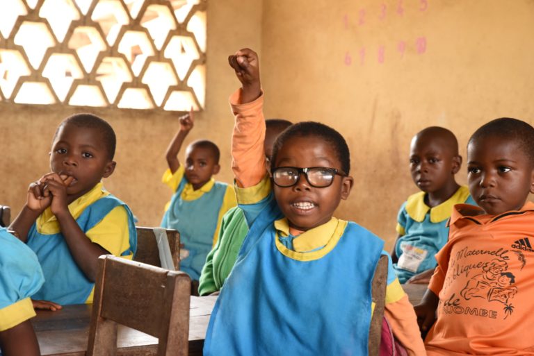 A boy wearing glasses raising his hand in a class at school.