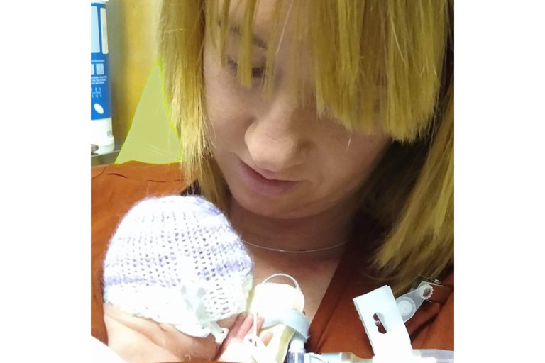 Liz is holding her baby Josie, who is very small, is wearing a knitted cap and has a number of tubes attached, against her chest and is looking down at her tenderly