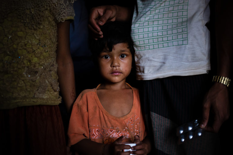 “A young girl wearing an orange shirt and holding a box of medicine is staring at the camera. Behind her are two adults, the one on her left is holding a packet of medicine.”