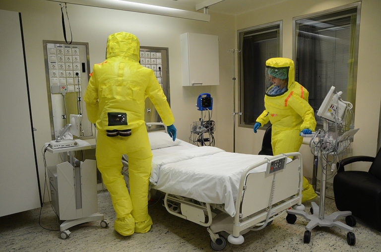 Inside a hospital room, two workers are dressed in protective gear for handling a patient with ebola virus