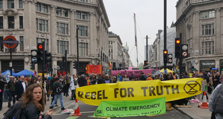 photo showing Extinction Rebellion protest in London, 2019. large yellow banner stating 'Rebel for truth'. Pink boat in the background with the text 'Tell the truth'