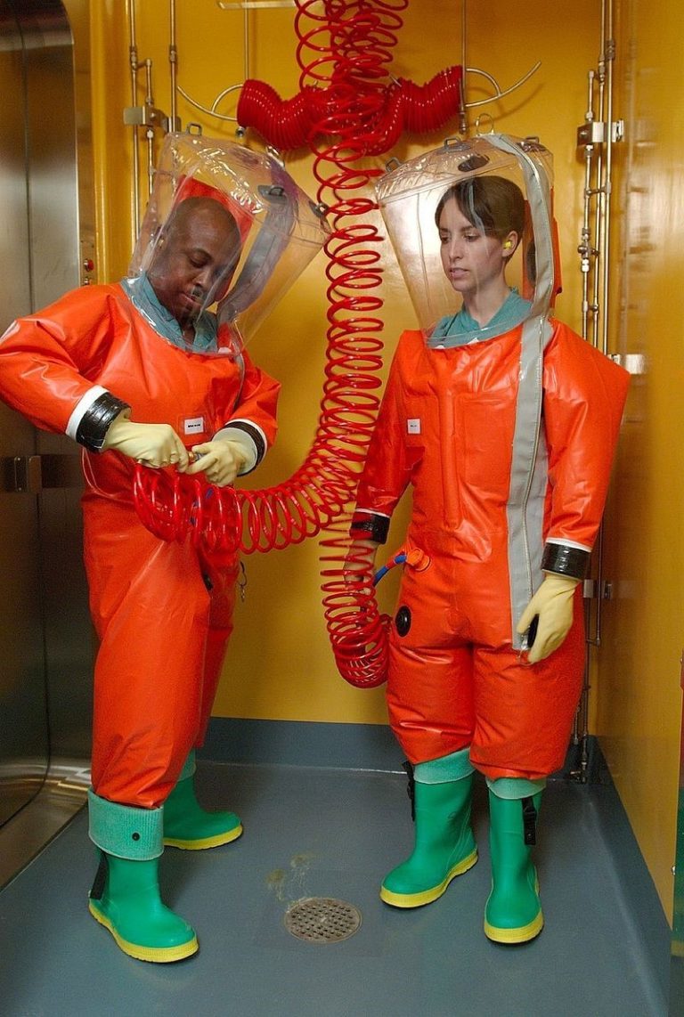 Two microbiologists putting on positive pressure biosafety suits which cover their entire body as well as a large clear cover over their head and wellington boots