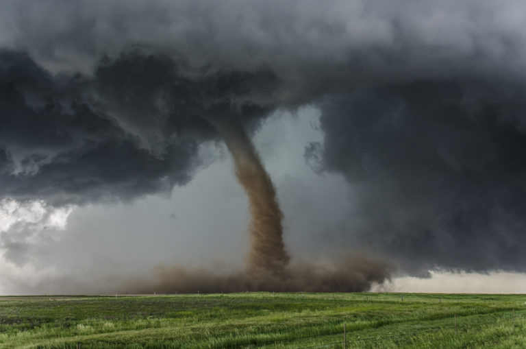 Tornado, centred in the image, coming from dark grey clouds and moving across green grass fields.