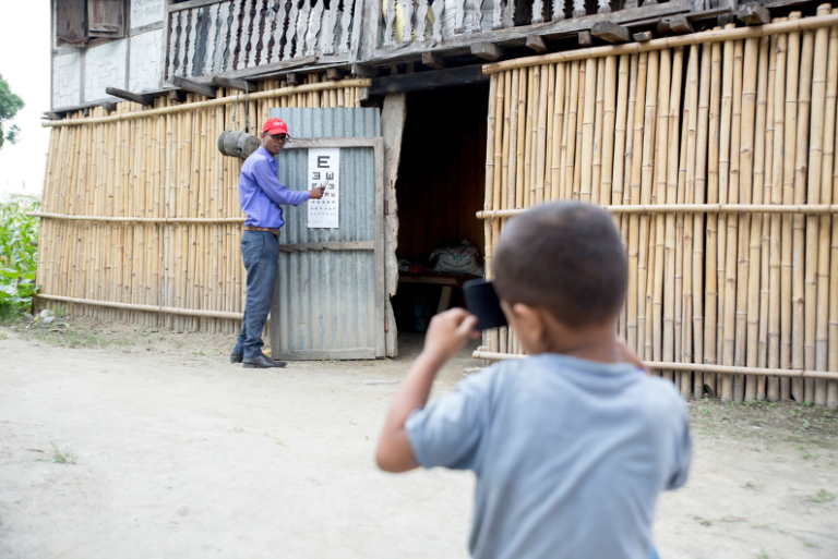 Health worker points to vision chart on the outside of a house made of bamboo, whilst boy is tested in the foreground.