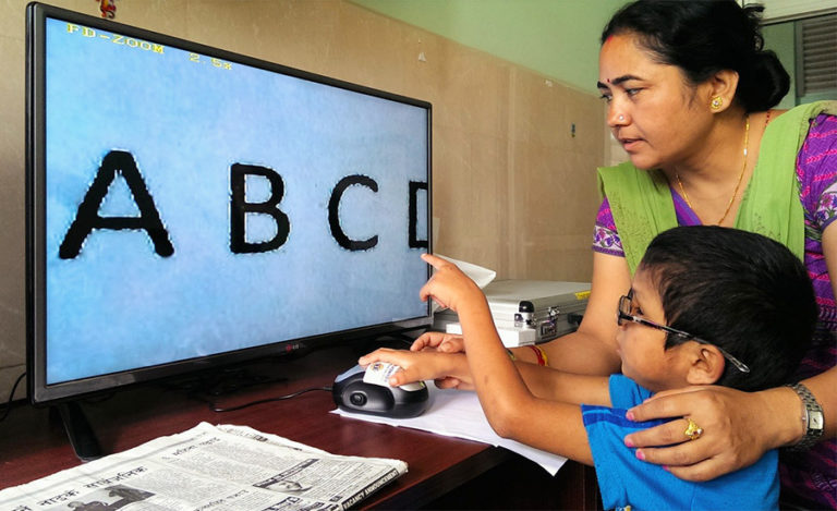 Small boy wearing spectacles is sitting at a desk and pointing at the magnified letters ABCD on a large computer screen whilst a woman operates the computer mouse standing behind him