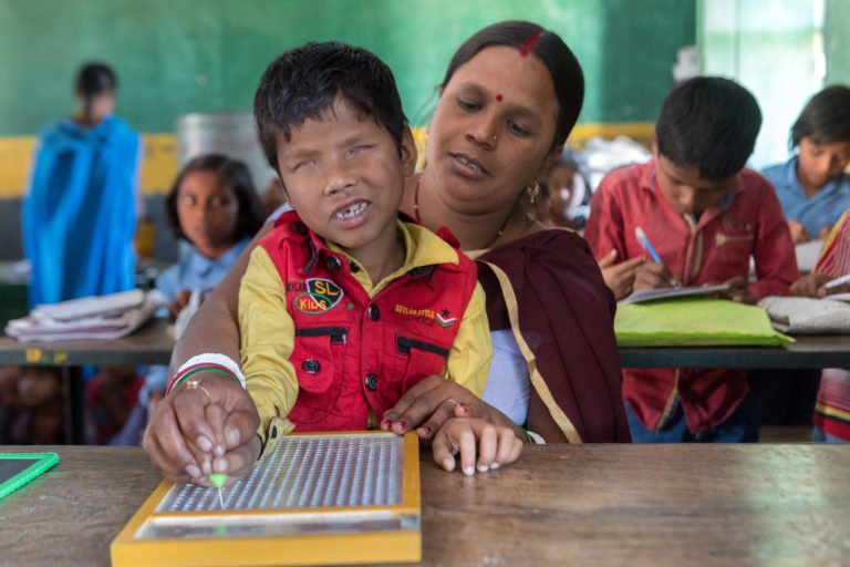 A boy without retina is sitting on the lap of a woman in school. She is helping him read a braille tablet