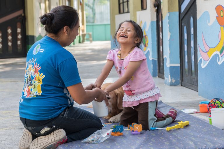 A girl laughing as she puts her hand into a bucket being given to her by a therapist.