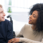 A woman smiling and talking to a young boy in a wheelchair.