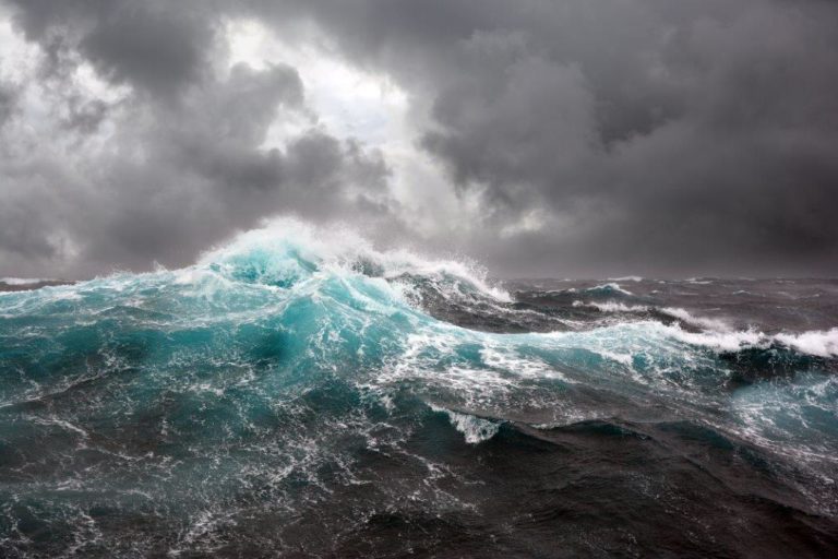 Colour enhanced photograph of a turbulent sea with waves and white crests, against grey clouds with a hint of blue sky through a break in the clouds