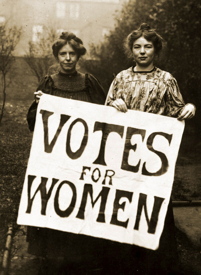 A black and white photgraph of Annie Kenney and Christabel Pankhurst holding a banner which reads 'VOTES FOR WOMEN'