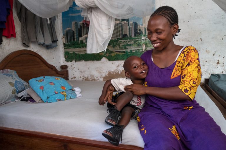 A boy is curling up to his mother and smiling as they sit on a bed.