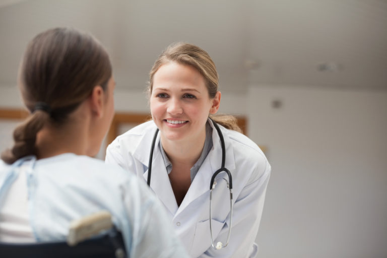 A smiling doctor bends down to be at eye level with her client in a wheelchair