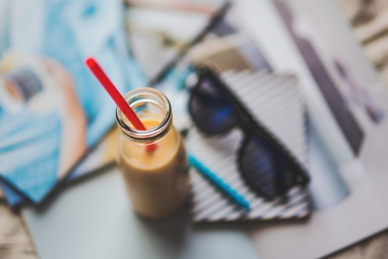 overhead view of a bottle of milky drink with a straw, sunglasses and notebook in the background