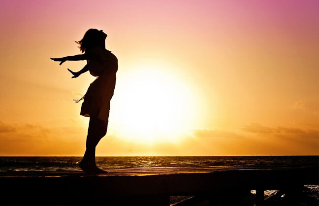 Image shows a silhouette of a person on a beach, looking up to the sky at sunset. 