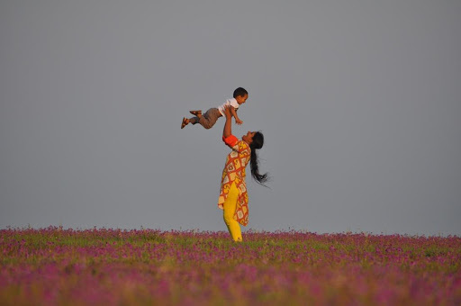 A colour photograph of a mother, wearing a yellow and orange shalwar kameez, lifting her child high above her head playfully