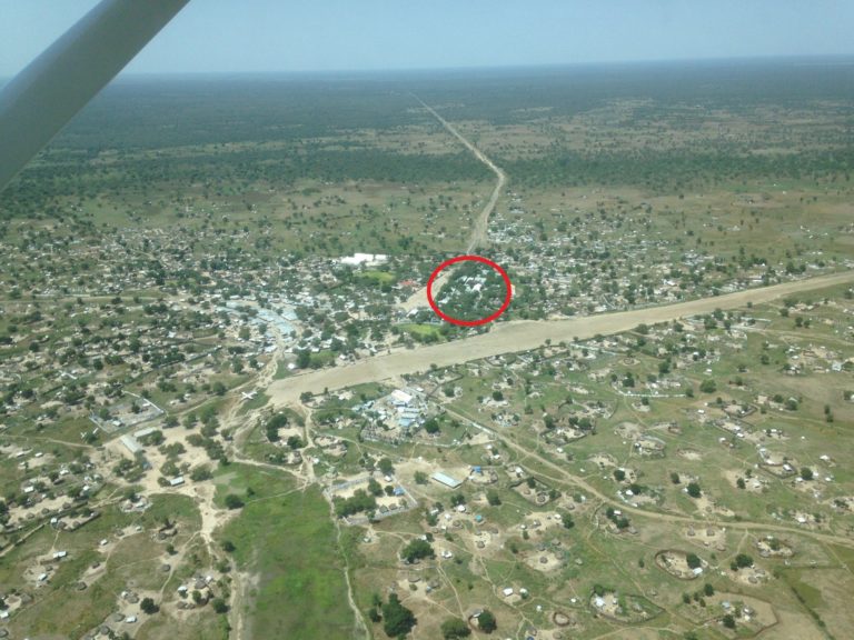 An aerial view of Lankien in South Sudan. There is a red circle showing where the hospital is. The landscape is mostly flat and green (but barren) with some buildings. There is an airport runway with an airplane on the tarmac. There is a forest in the distance.