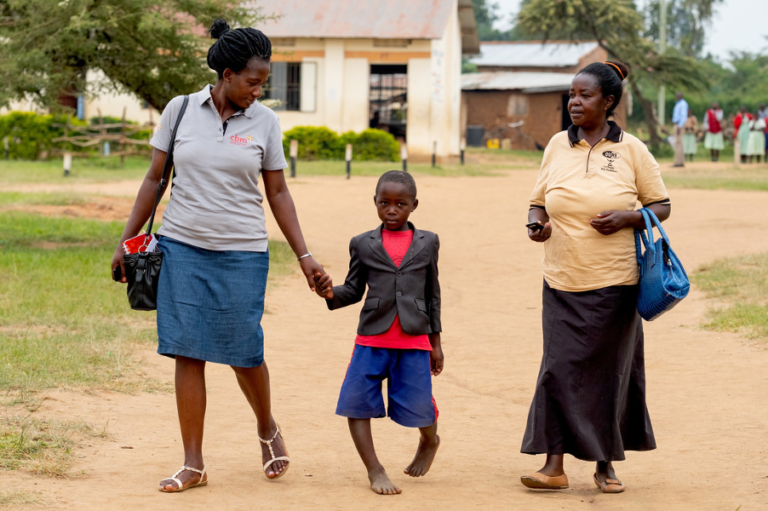 Young boy with severe bowlegs walking between two community health workers outside the school he attends in Rwemirabyo, Uganda