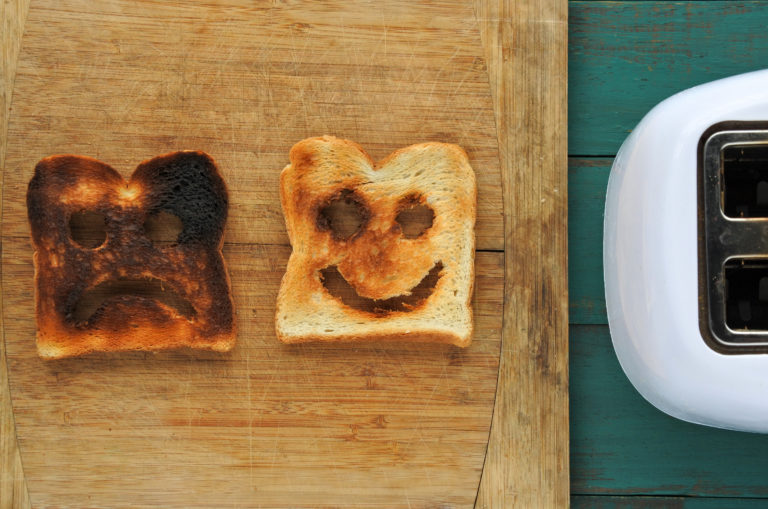 two pieces of toast on a table by a toaster, one black and dark brown (burned) with a sad face carved into it, one paler brown and white with a happy face carved into it