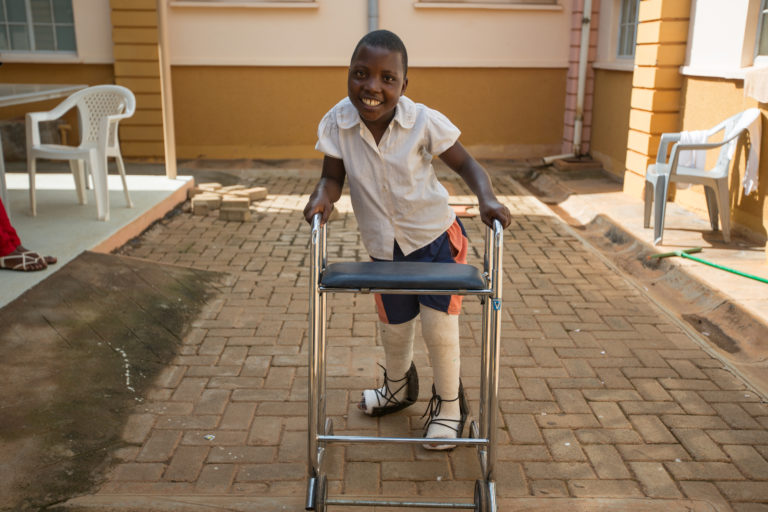 A girl with casts on her legs is using a walking frame. She is smiling as she walks towards the camera