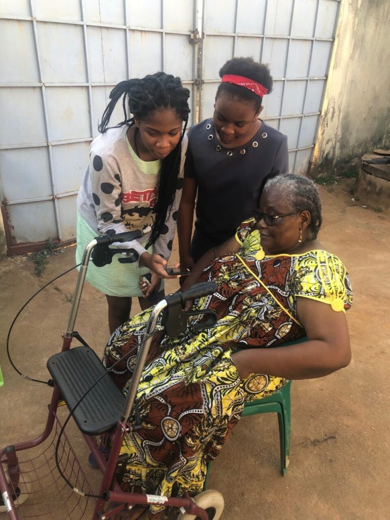 Three people are shown in the photo - a Cameroonian grandmother and her two female grandchildren. She is in a wheelchair and her grandchildren are looking at something on her phone, helping her update an app
