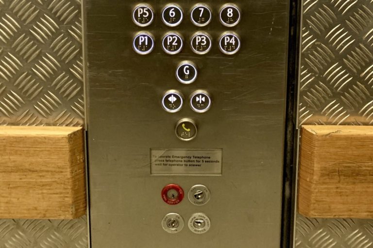 An image of a lift control panel with the ground floor separate, the phone button protected by a ridge, and a series of key holes underneath.