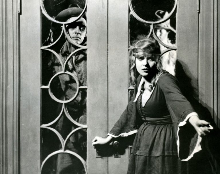 Still image from the black and white film The Warrens of Virginia, where a woman stands in front of a closed door with a man peering through from the other side