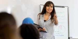 Teacher stands at a classroom whiteboard and points towards a student, smiling